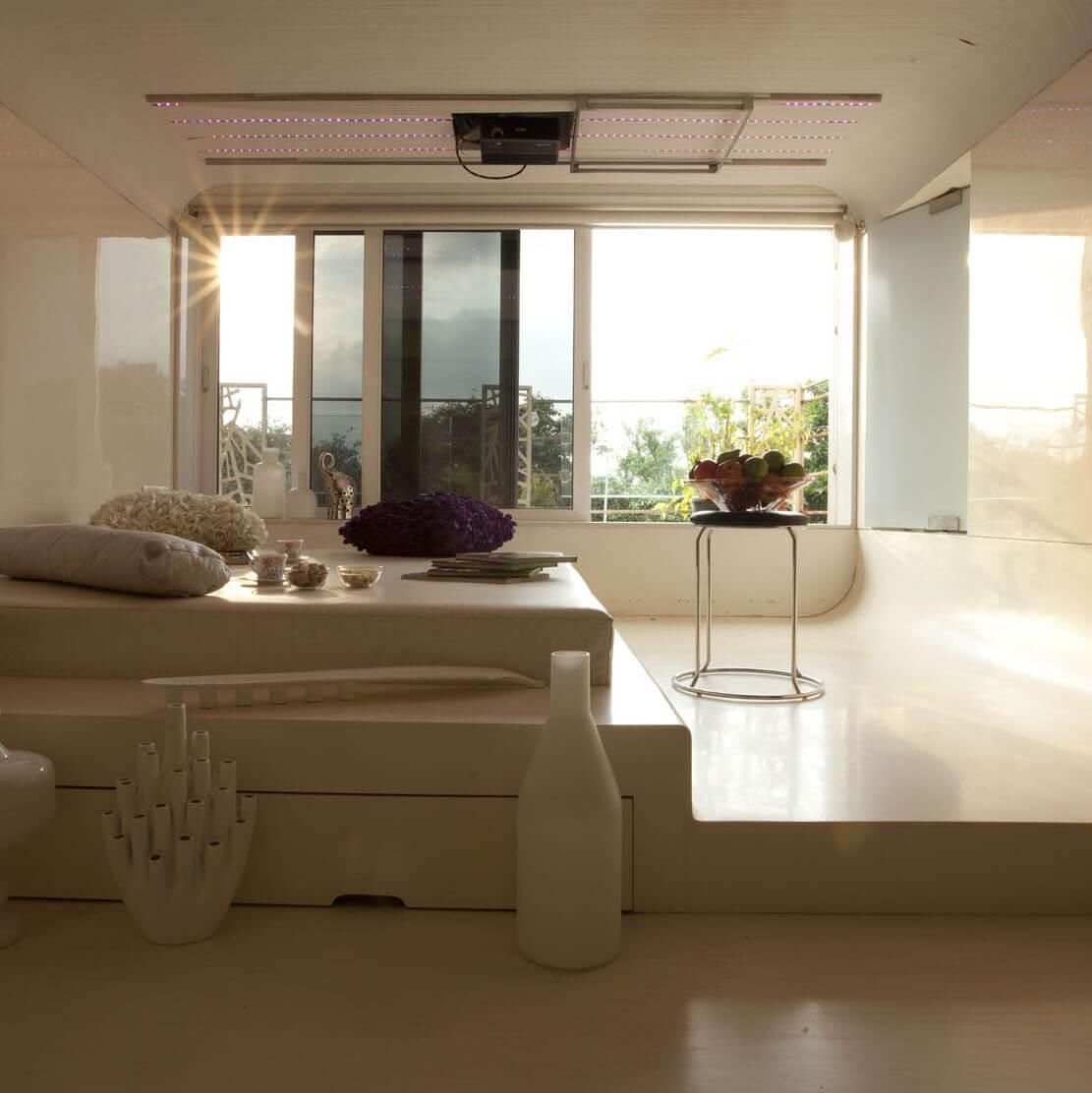 Modern minimalist white room with raised platform, cushions, and large sunlit windows.