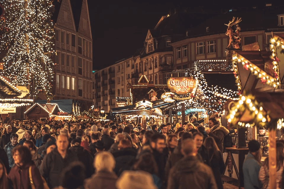 A bustling Christmas market at night, filled with people, festive lights, and decorated stalls.