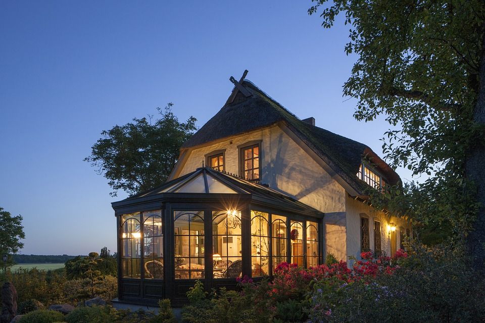 Charming cottage with a lit glass sunroom at dusk, casting a warm glow.