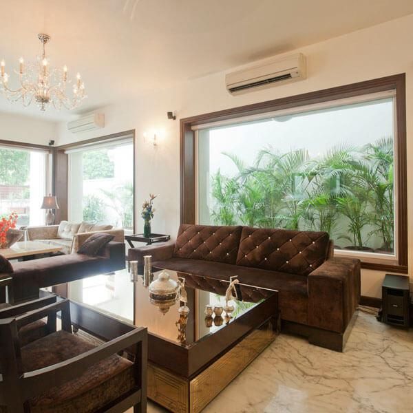 Modern living room: brown sofa, glass coffee table, and large windows revealing green foliage.
