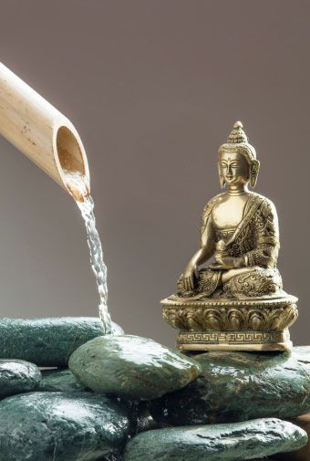 Golden Buddha statue meditating beside water flowing from a bamboo spout onto dark stones.
