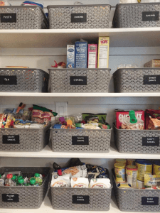 An organized pantry features white shelves filled with labeled gray storage baskets containing various food items.