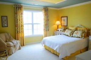 A bright yellow bedroom with a bed, white bedding, floral curtains, and a large window.
