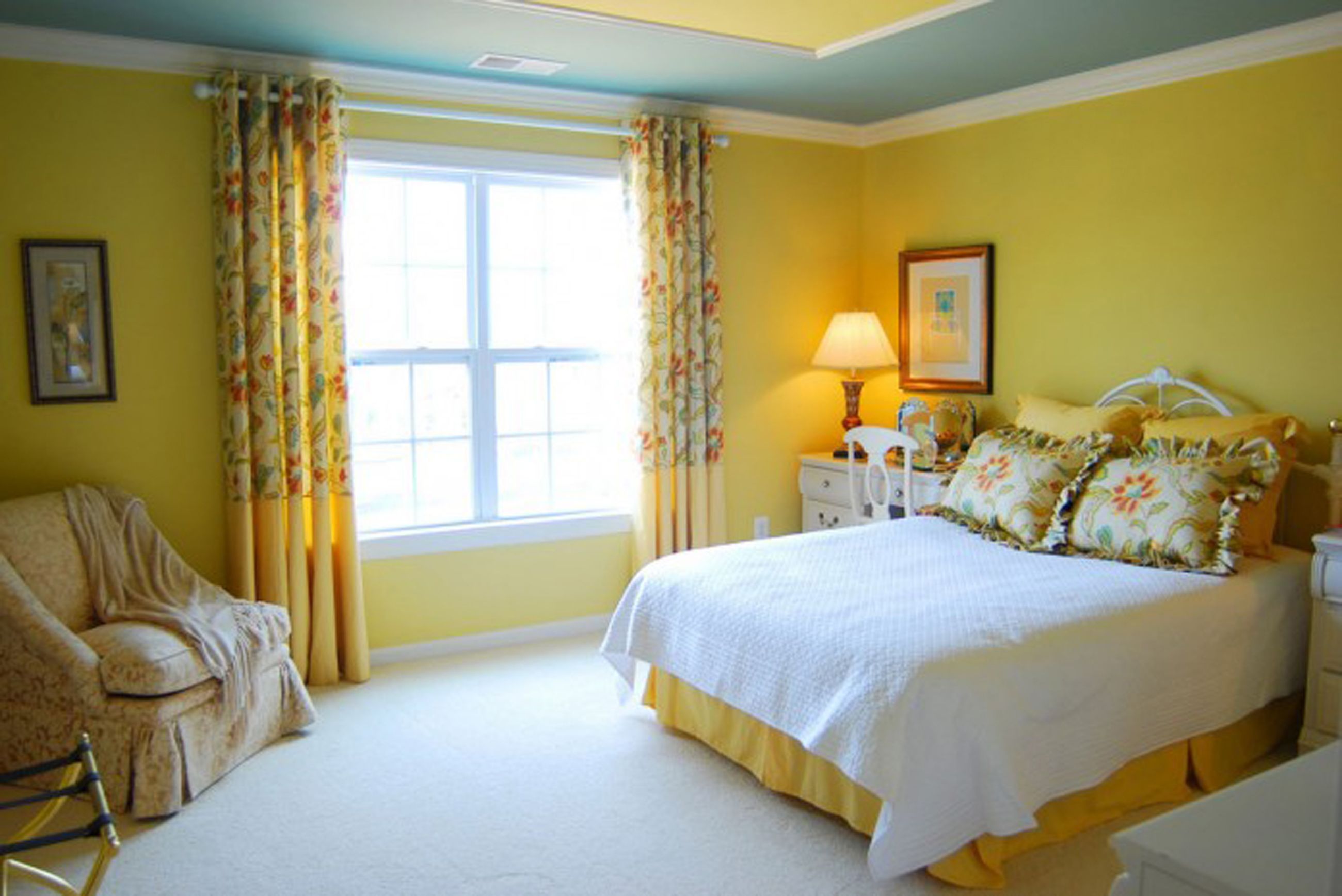 A bright yellow bedroom with a white bed, floral curtains, and a comfortable armchair.