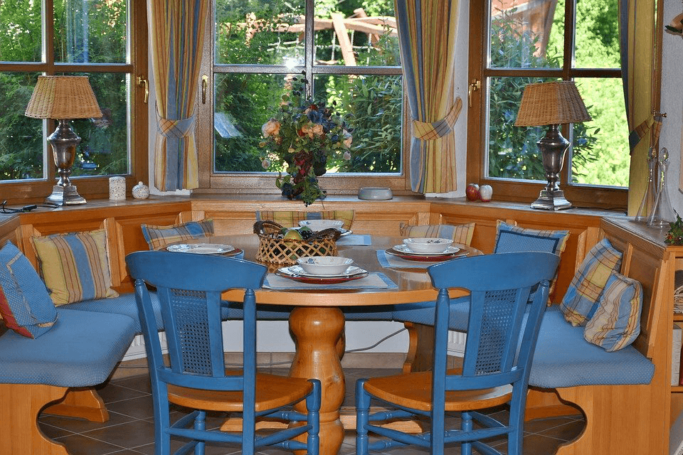 Cozy dining nook with a round wooden table, blue benches, and chairs by windows.
