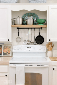 White kitchen featuring an electric stove, wooden shelf, and hanging cooking utensils.