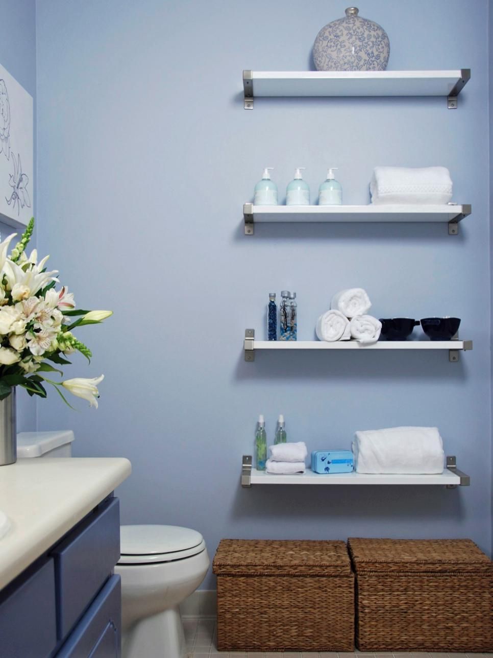 Light blue bathroom with white floating shelves, organized towels, toiletries, and a floral arrangement.