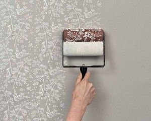 Hand applies a light botanical pattern to a gray wall using a decorative paint roller.