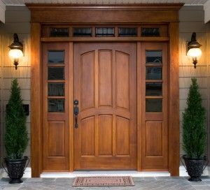 Elegant wooden front door with side windows, transom, lights, and potted evergreens.