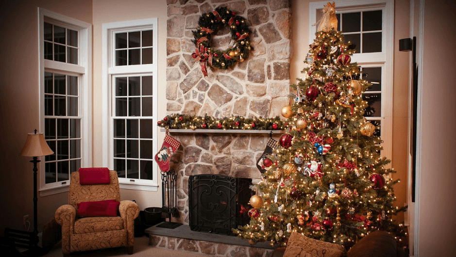 Lit Christmas tree, stone fireplace with wreath, and cozy armchair in a festive room.