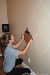 Woman crouching, carefully applying an ornate Moroccan stencil pattern to a beige wall.