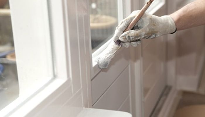 Gloved hand painting a white window frame with a paintbrush, applying fresh white paint.