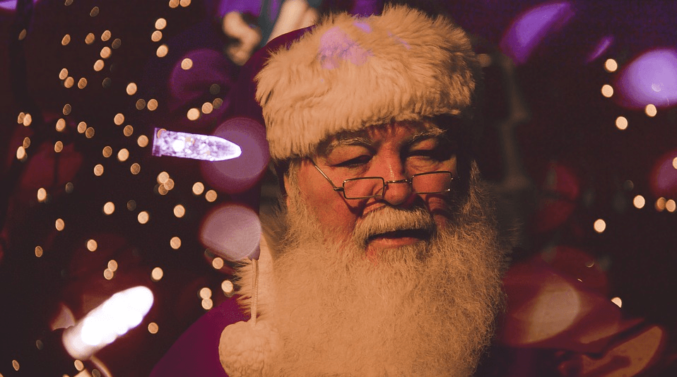 Santa Claus with white beard, red hat, glasses, and closed eyes, against blurred festive lights.
