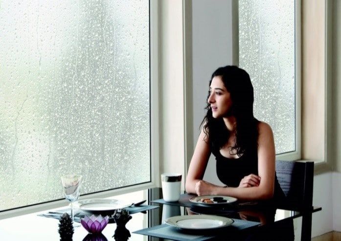 A woman looks thoughtfully out a large window at a rainy scene from a dining table.