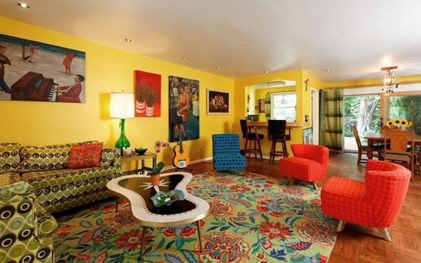 Vibrant retro living room with yellow walls, floral sofa, red and blue chairs, and a colorful rug.