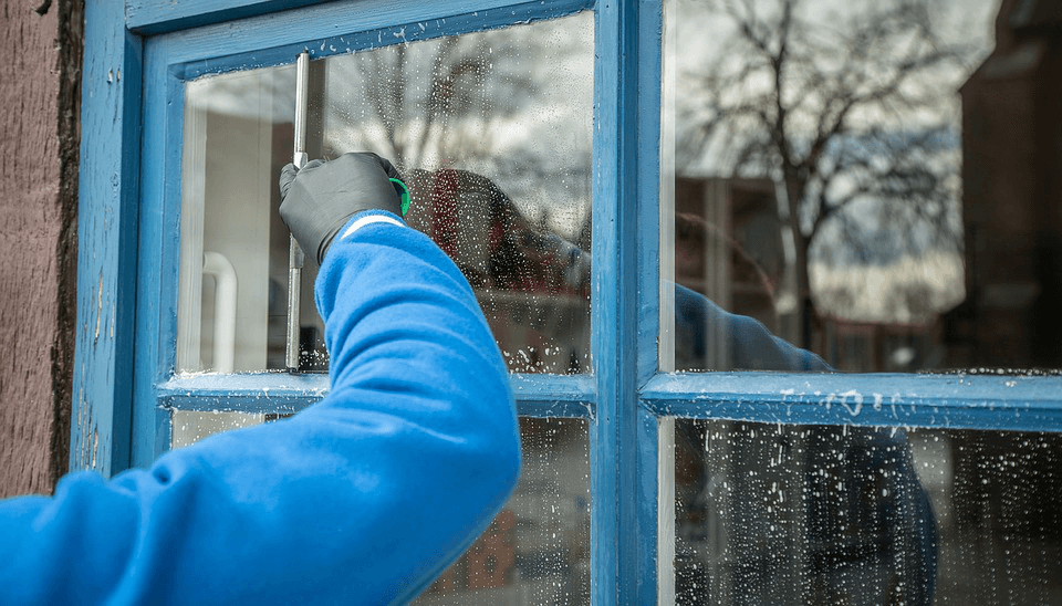 Person in blue jacket cleans a blue-framed window with a squeegee.