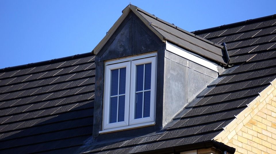 White dormer window with glass panes on a dark tiled roof against a blue sky.