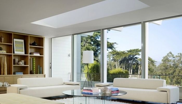 Modern living room with white sofas, glass table, large windows, and outdoor trees.